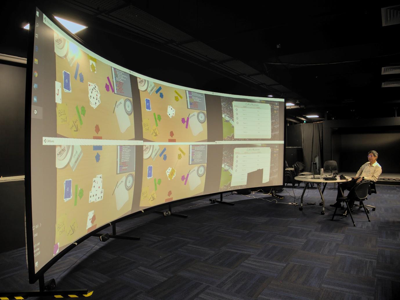 An instructor sits at a desk facing a projector screen showing 4 CAVRoom class sessions and 2 other screens