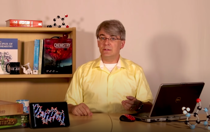 A person sitting behind a desk and speaking to the viewer. Around them on the desk and a shelf behind them are a laptop computer, and various chemistry textbooks and molecular models.